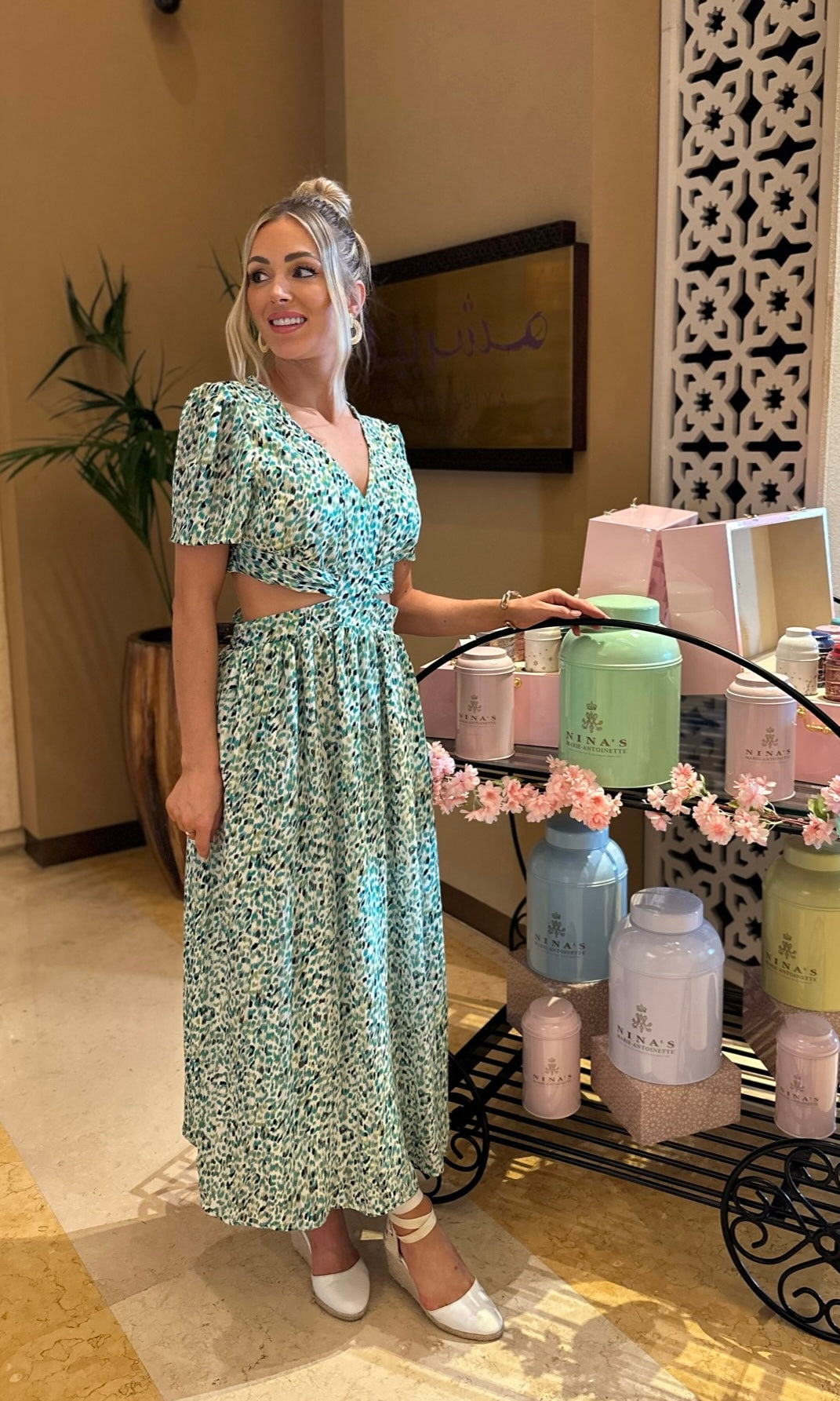 Woman in a floral dress standing in a room with decorative items and plants.