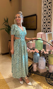 Woman in a floral dress standing in a room with decorative items and plants.